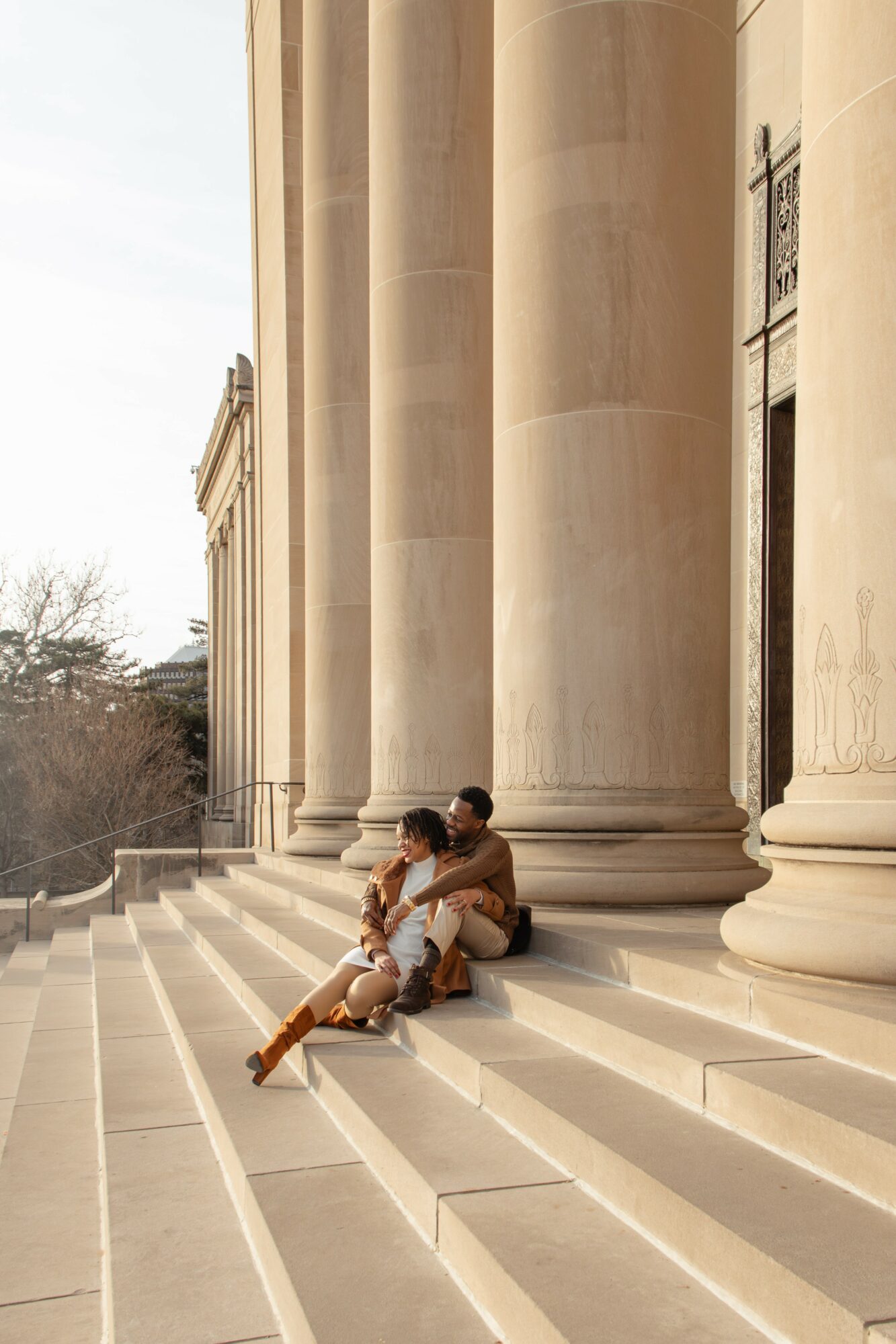 Two people sit on steps in front of large columns of a building, with trees in the background.