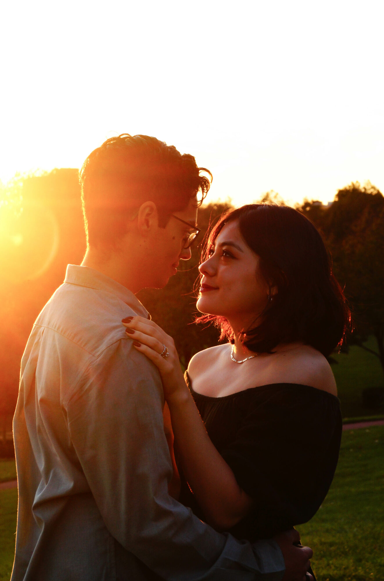 A couple stands close together outdoors during sunset, gazing at each other with trees in the background.