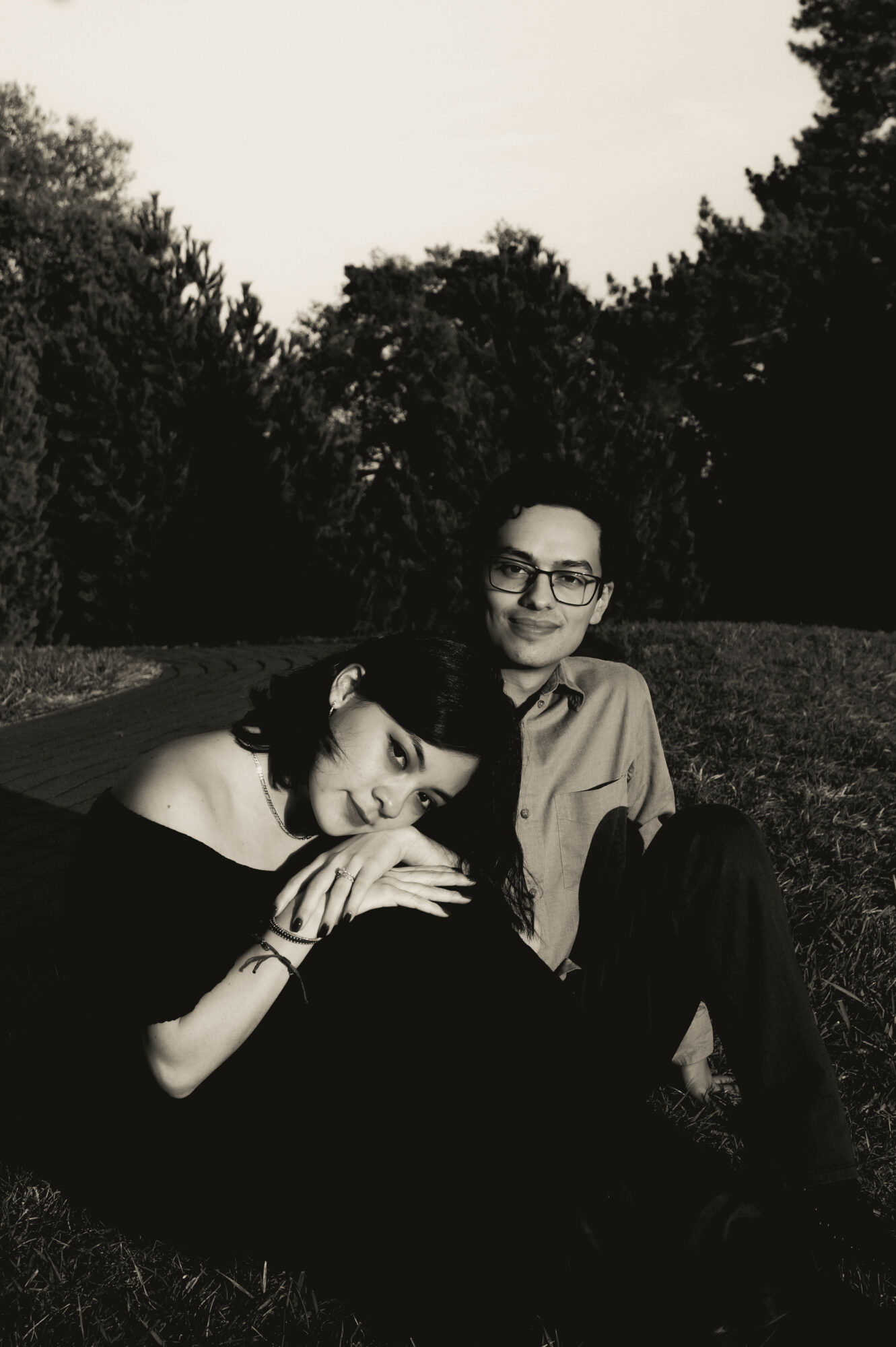 Two people sitting on grass outdoors, one resting head on the other's lap, trees in background, daytime.