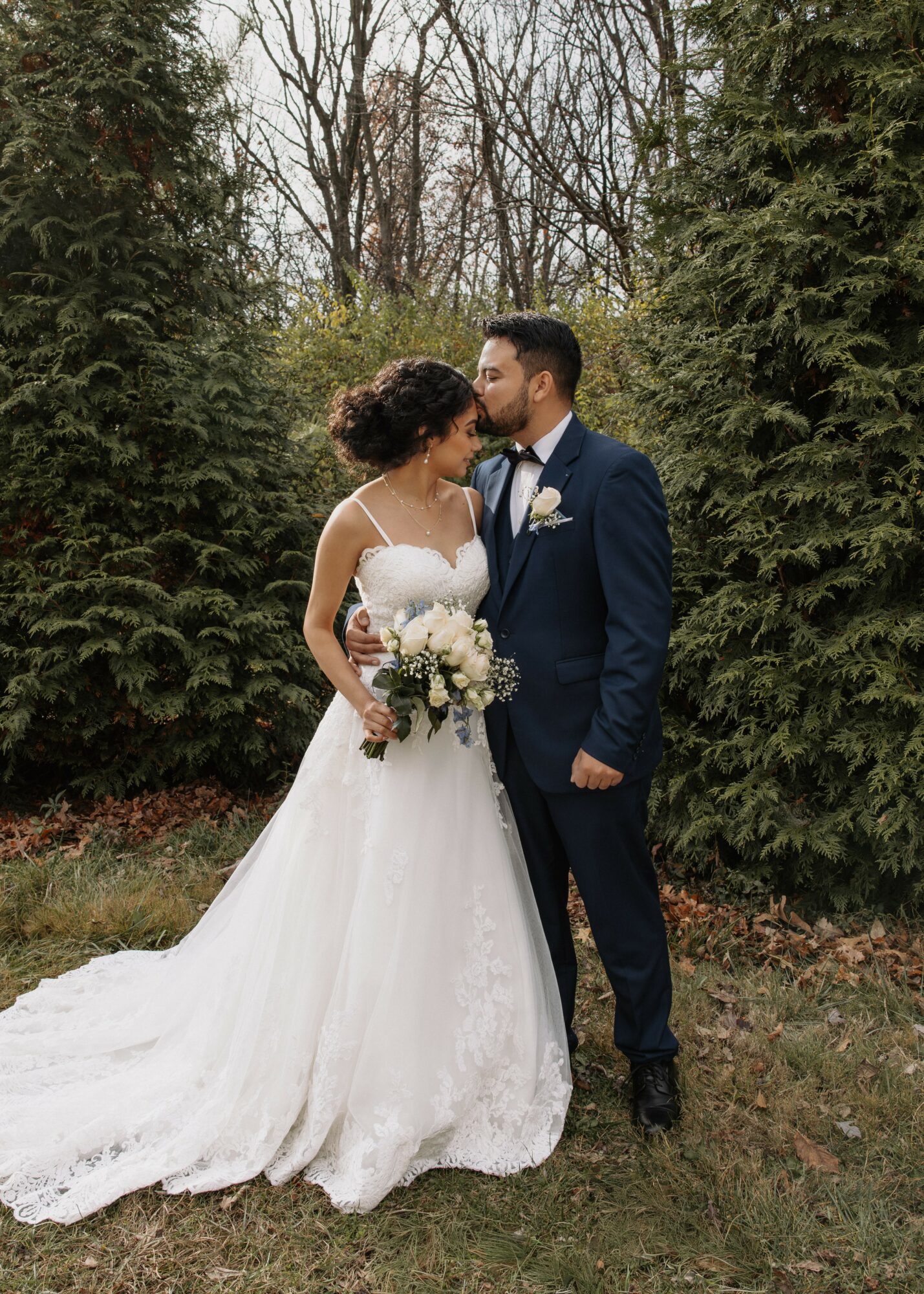 Bride and groom stand close, surrounded by trees, gazing at each other, the bride holding a bouquet, outdoors.