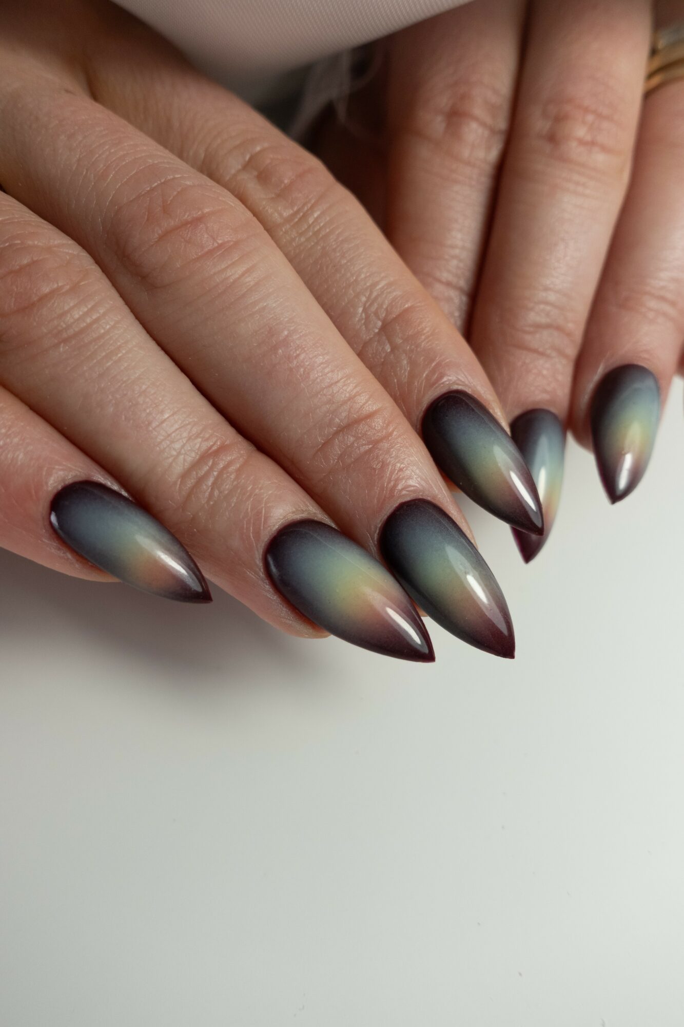 Close-up of hands with long, pointed nails painted in a metallic gradient color, against a white background.