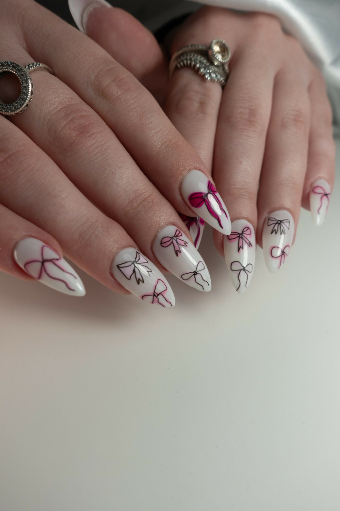 Two hands with decorated nails and rings, nails have white base with pink and black bow designs, on a white surface.