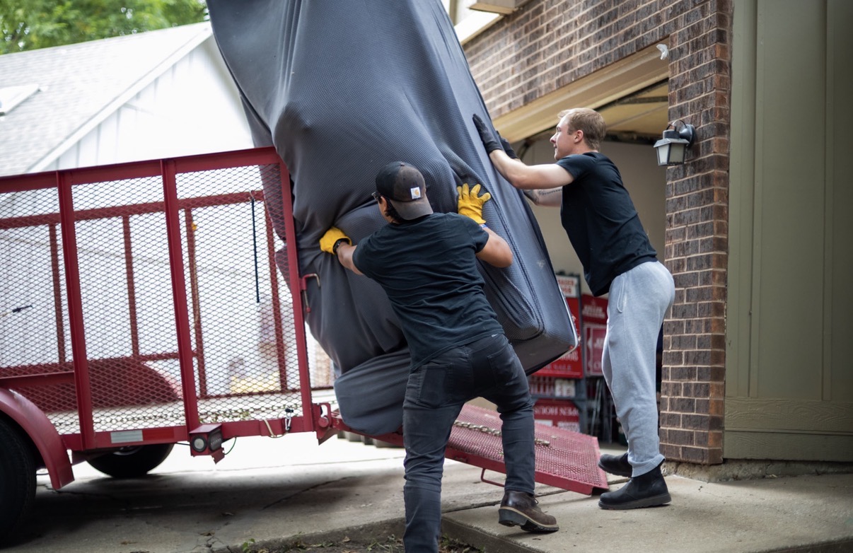 Two men are moving a large object into a building, with a red cart nearby. One man is pushing, the other is guiding.