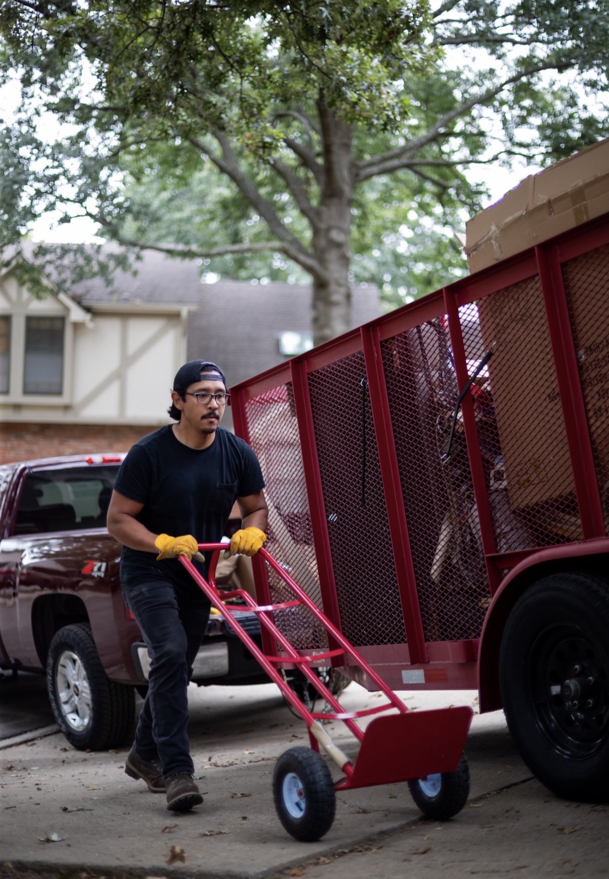 Person walking beside a red utility trailer attached to a vehicle, holding a pink hand truck with yellow gloves, outdoors with trees and houses.