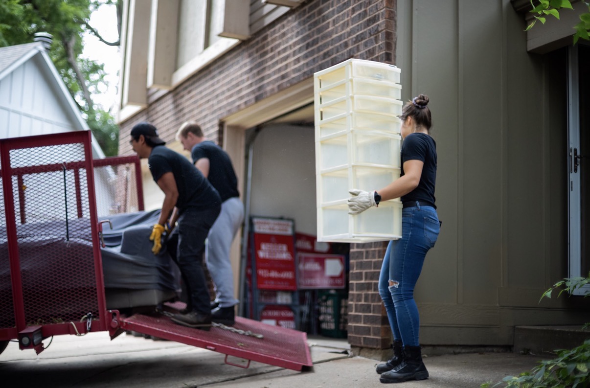 Three people unload a large plastic storage container from a truck outside a building.
