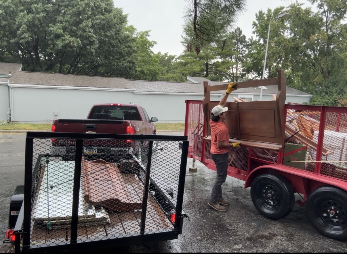 Person loading furniture onto a red trailer, with a black trailer nearby, outdoors with trees and a building in the background.