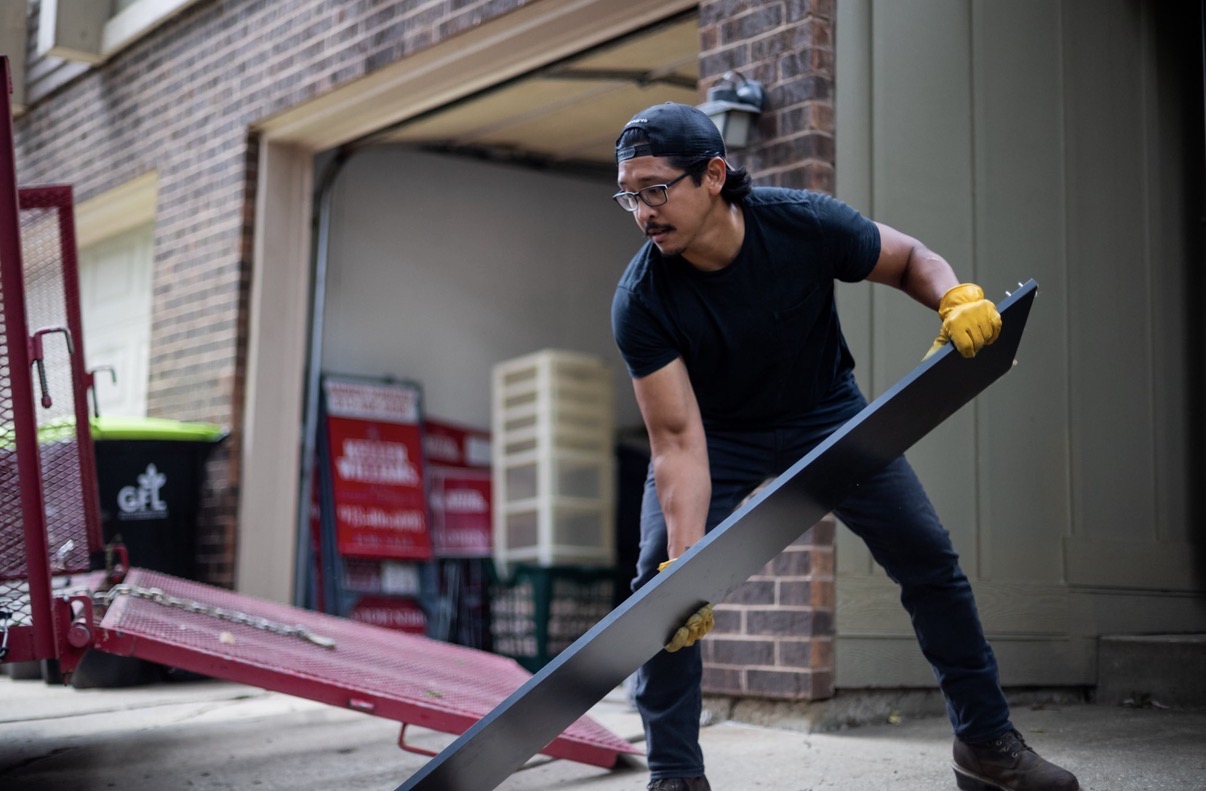 Person wearing glasses and yellow gloves lifting a large metal sheet outside a building.