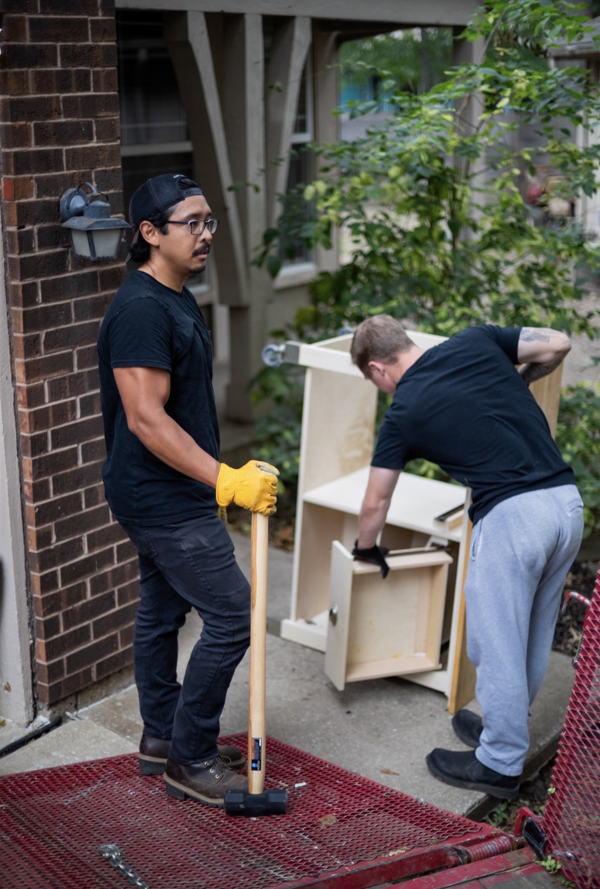 Two men working outdoors near a brick wall and green trees, one holding a hammer and the other assembling furniture.