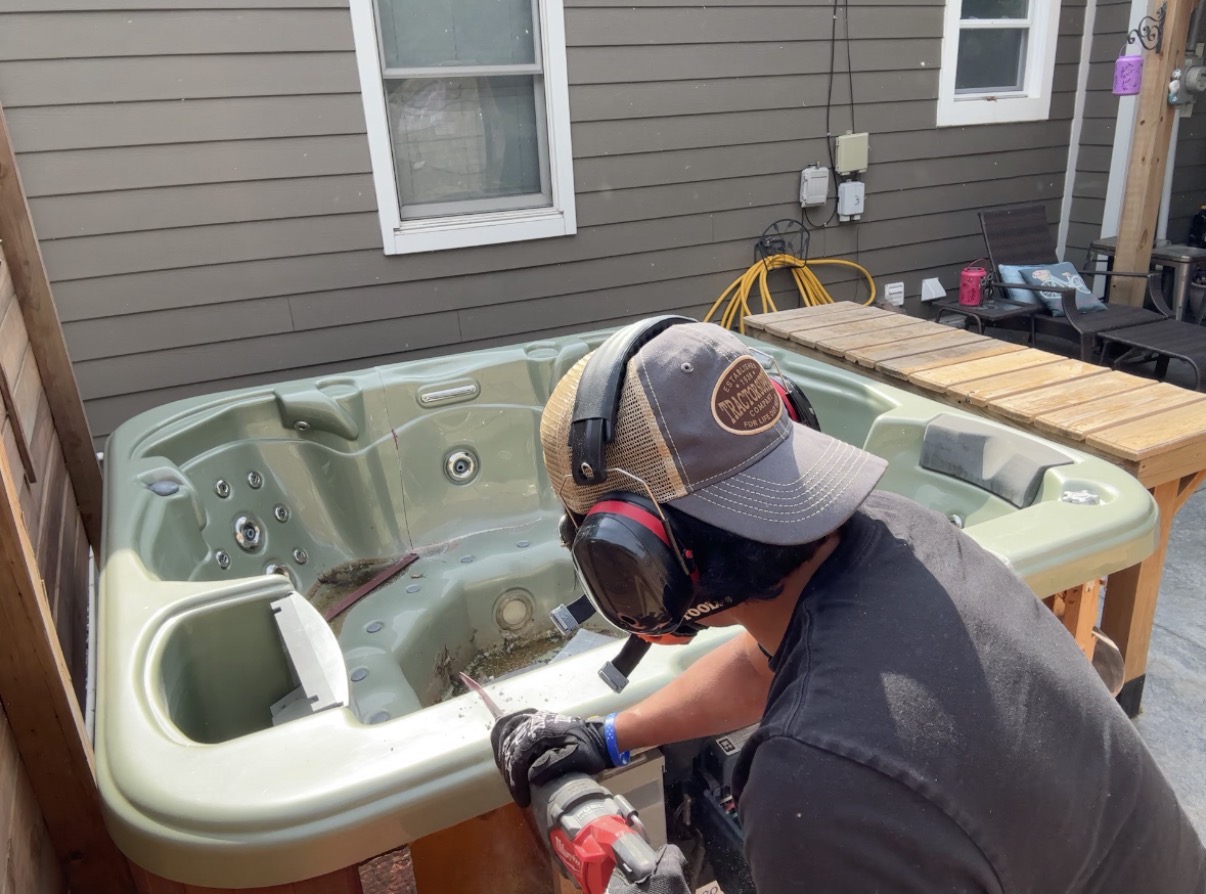 Person working on a hot tub outdoors, wearing a cap and headphones, with a wooden table nearby.