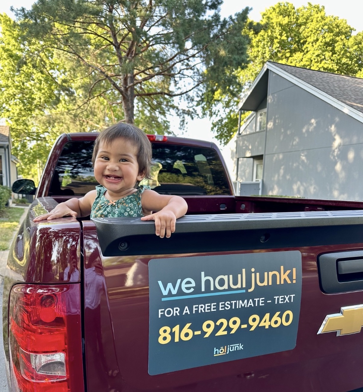 Child smiling in truck bed with trees and house in background, advertisement on truck side, text about junk removal service, phone number.