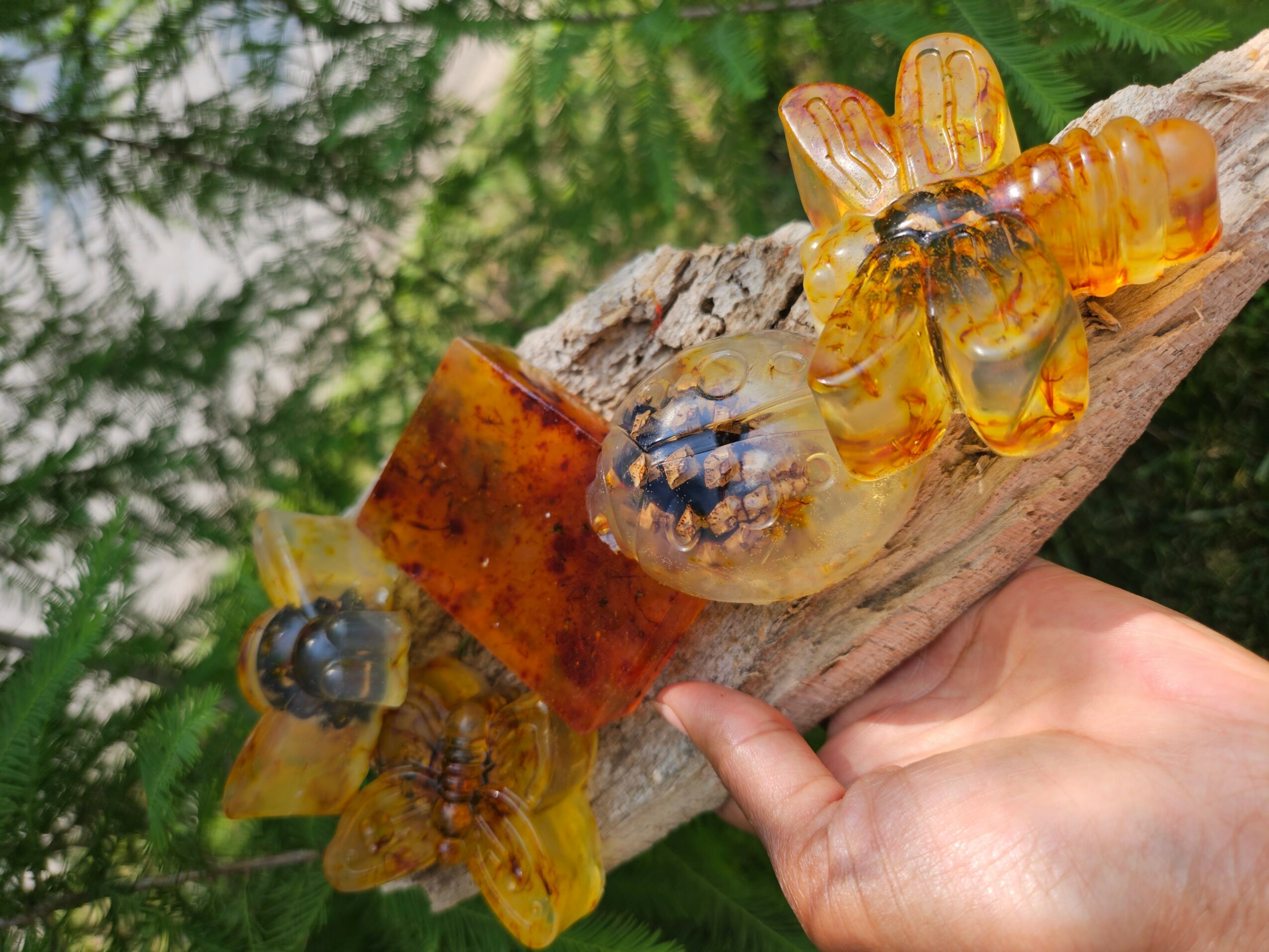 Hand holding a piece of wood with amber-colored bee-shaped ornaments on it, outdoors with green foliage background.