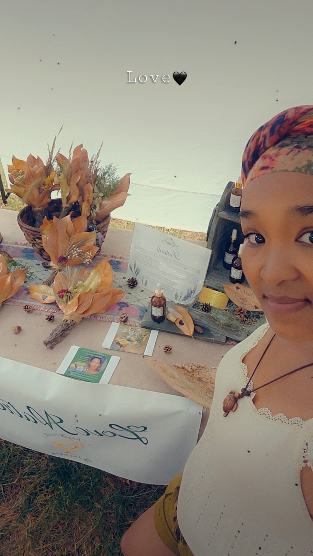 Person with colorful headscarf smiling at a table with flowers and a cake, outdoors, sky background.