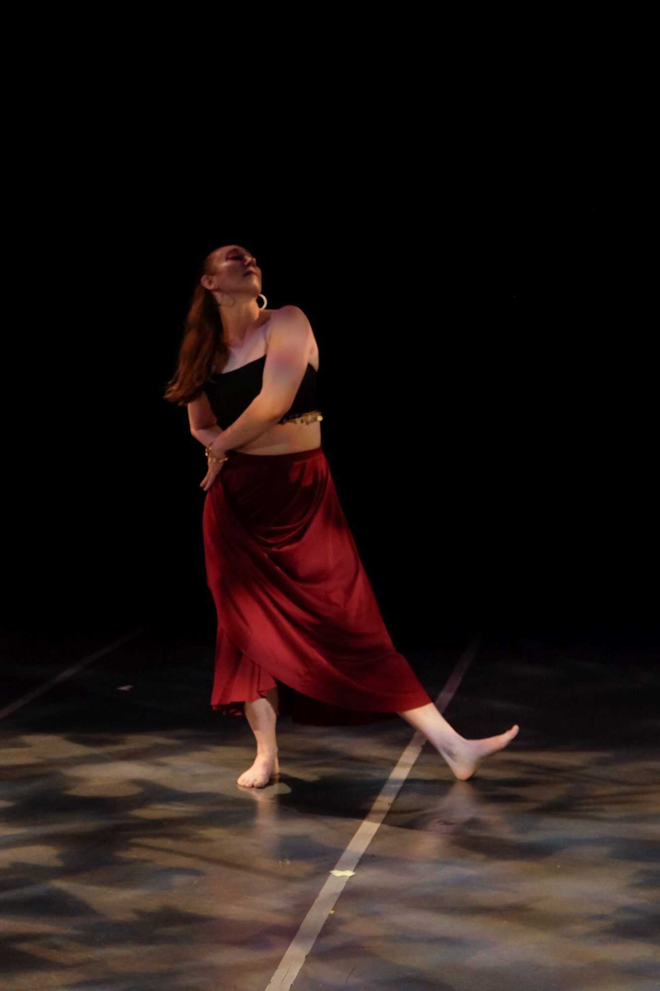 A woman dances barefoot on stage with a black background, wearing a black top and red skirt, in a dynamic pose.