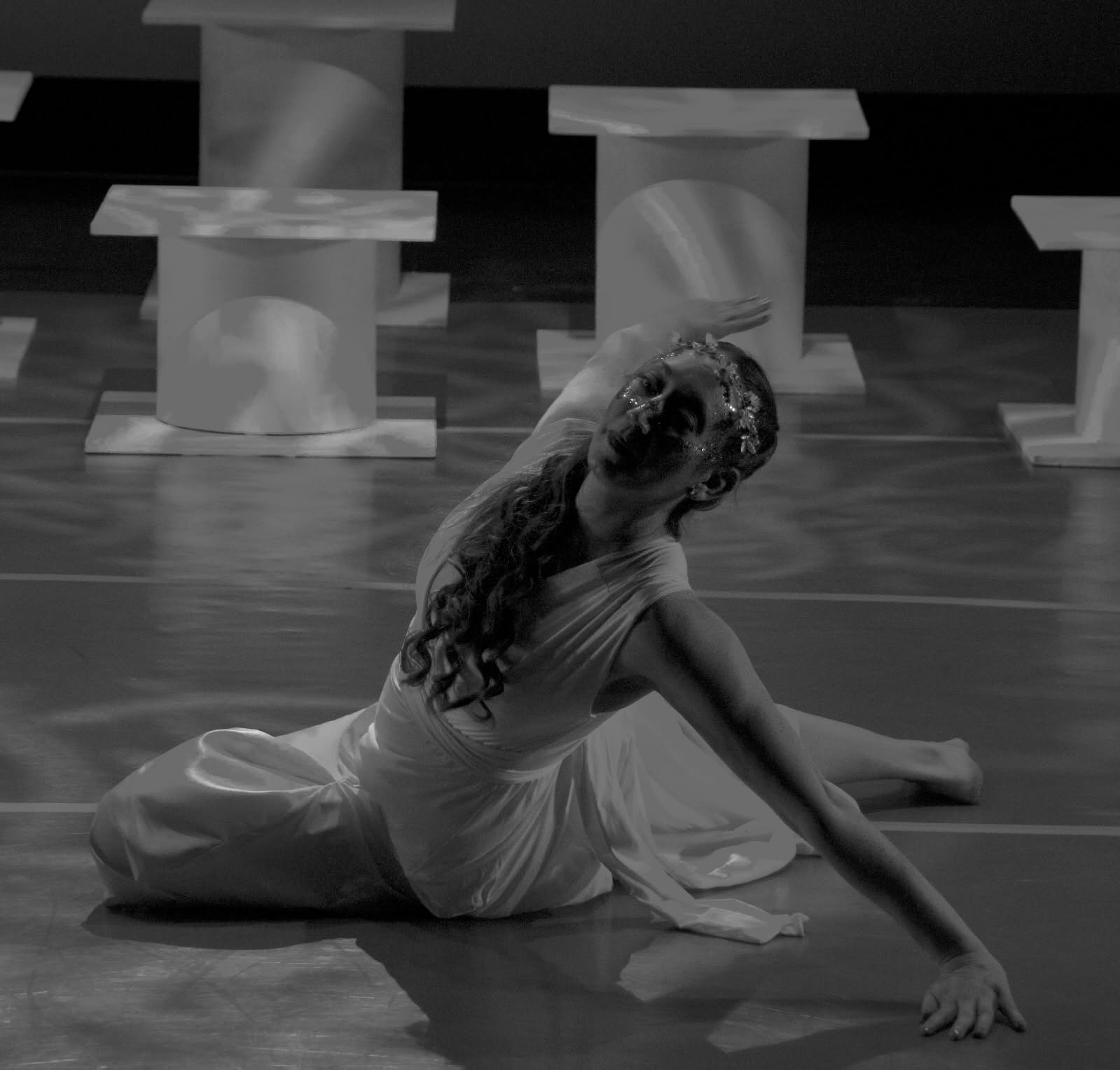 Person performing dance move on floor with props in background, in black and white.
