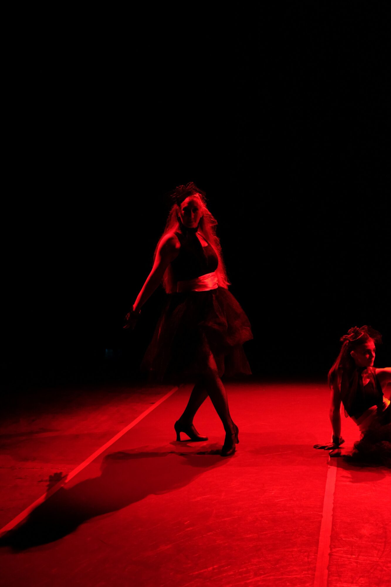 Two dancers perform on stage with red lighting, one standing and one kneeling, in a dark background.
