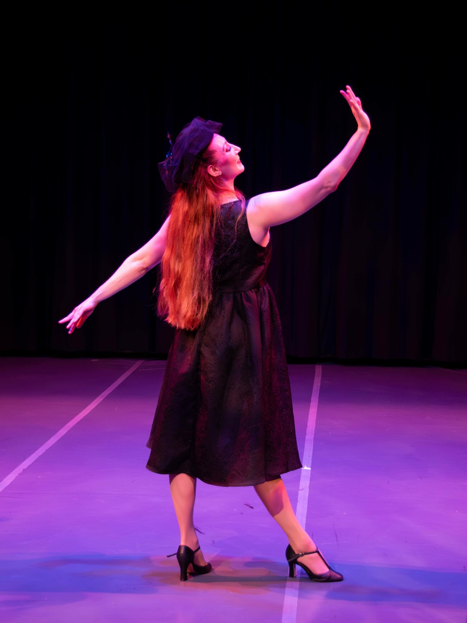 Woman with long red hair in a black dress and hat dancing on stage with one arm raised.
