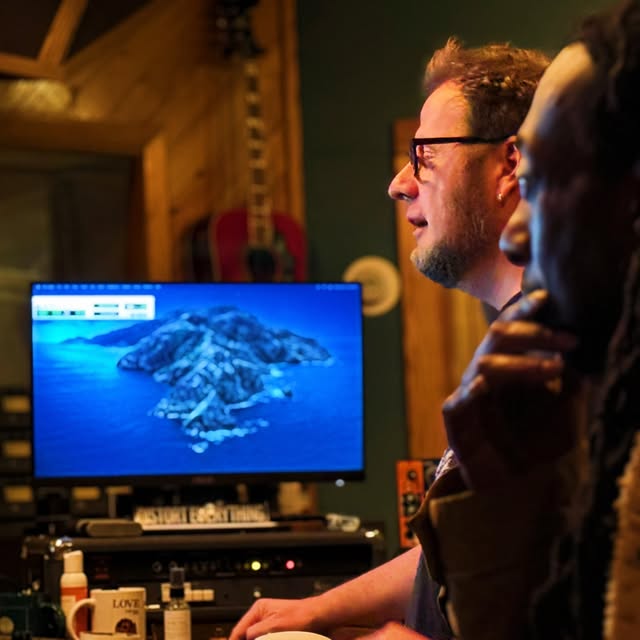 Two people watching a computer screen displaying an island surrounded by water in a wooden room.