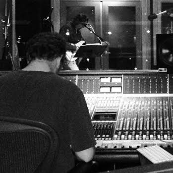 Person working at a large audio mixing console in a recording studio, with a singer at a microphone in the background.