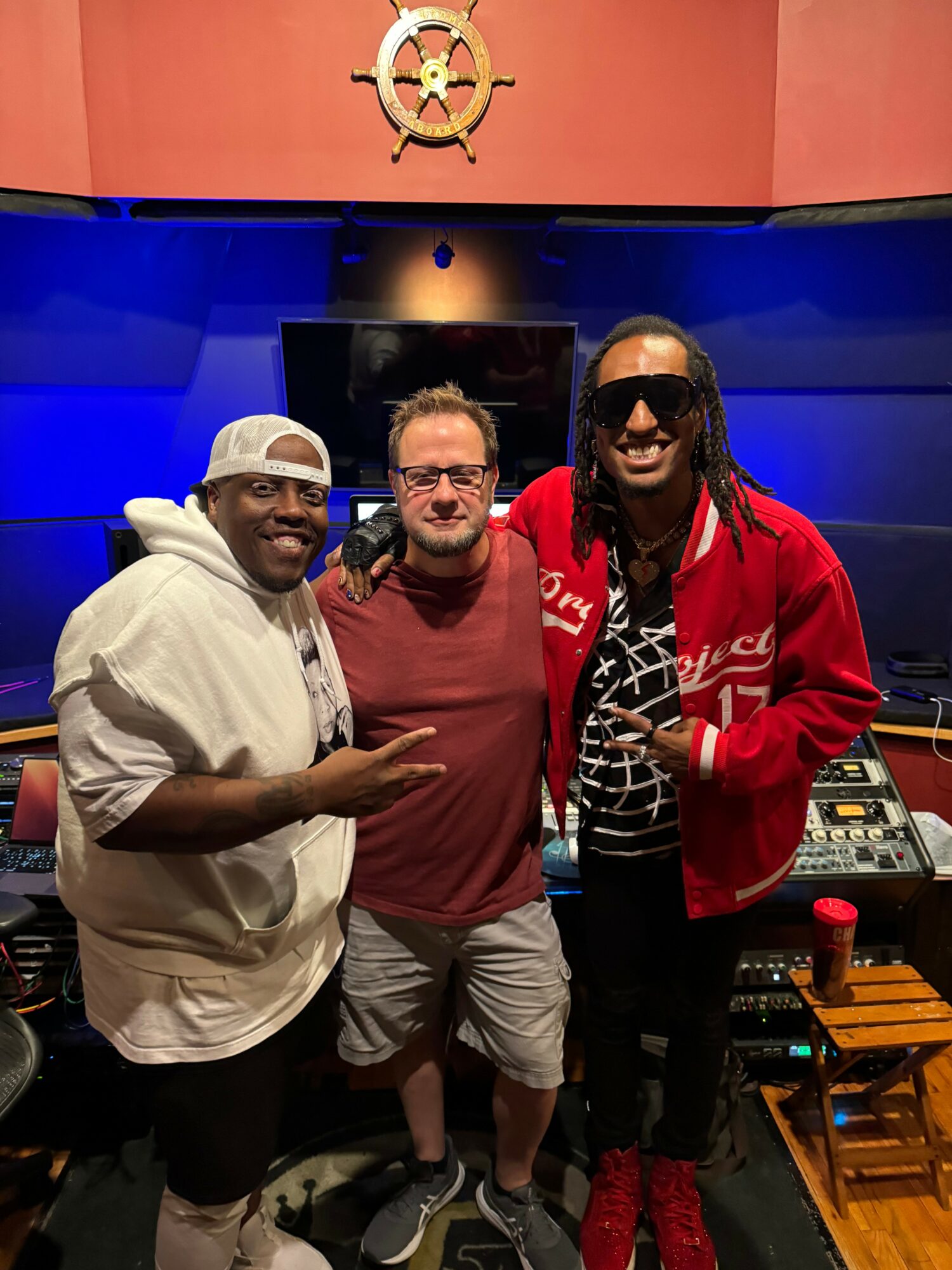 Three men standing together in a recording studio, smiling, with a nautical steering wheel decoration on the wall behind them.