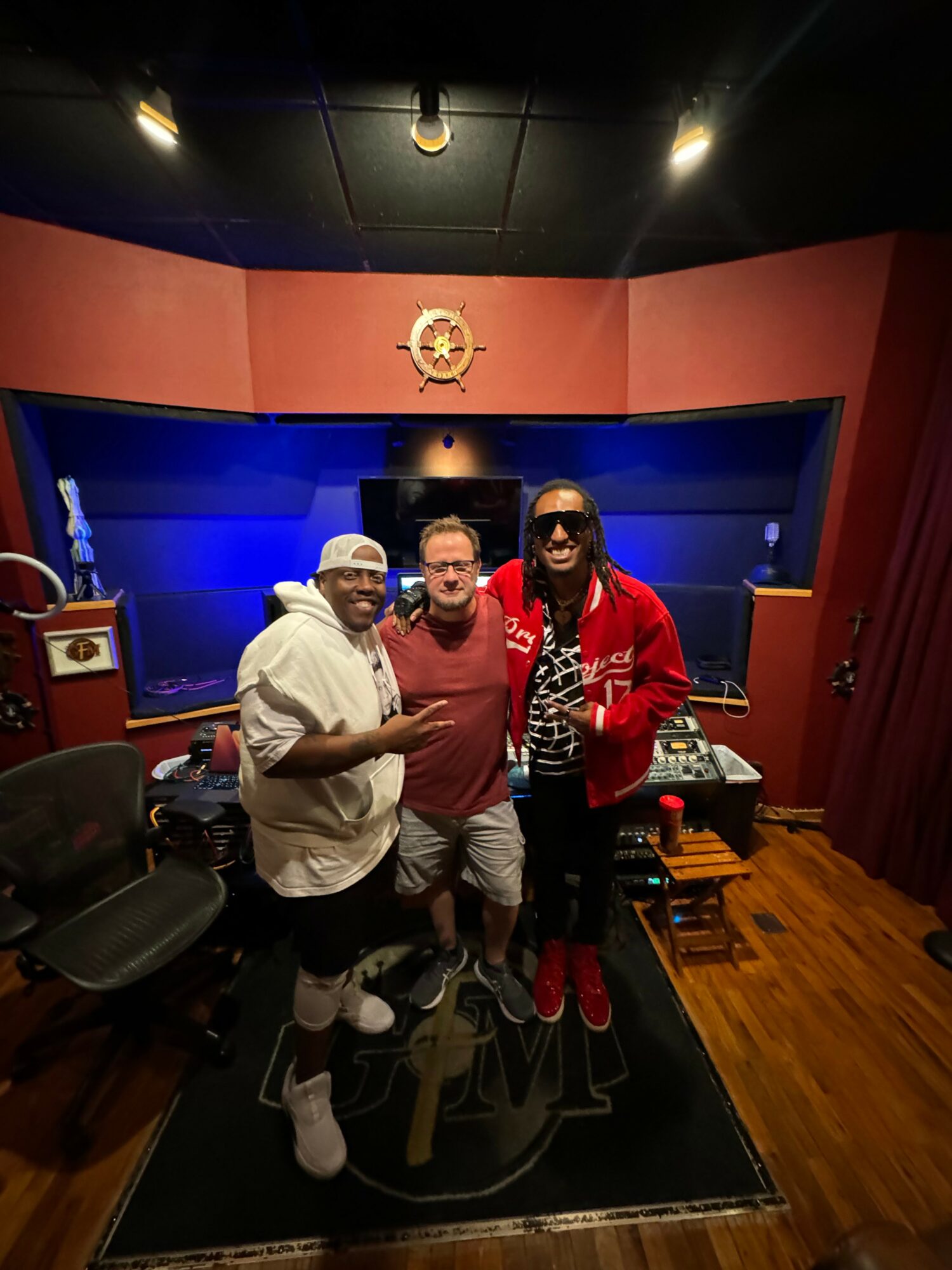 Three people standing in a recording studio with red and blue walls, a star emblem, and audio equipment.