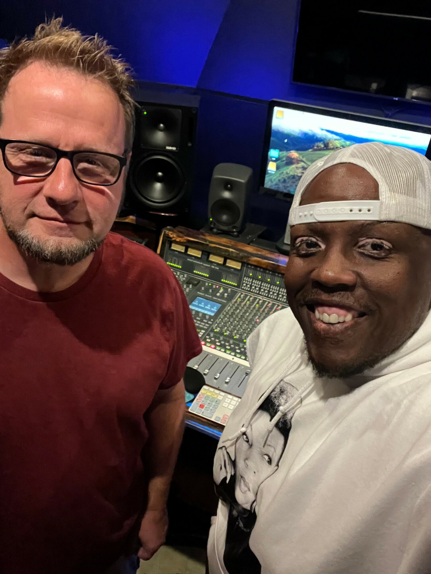 Two men in a recording studio, one wearing glasses and a maroon shirt, the other in a white shirt and cap, smiling.