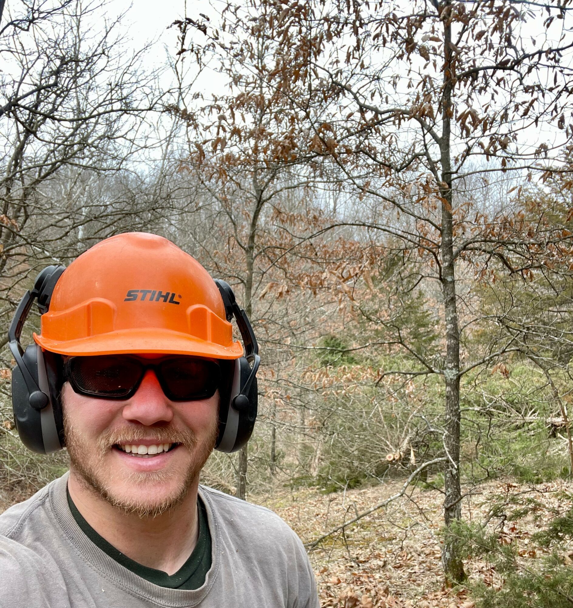 Person outdoors wearing orange helmet, sunglasses, and headphones, smiling, with trees and leafless branches in background.