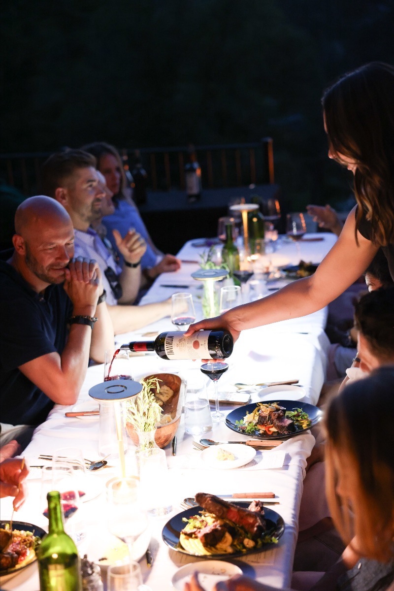 People dining outdoors at night, with a woman serving food to a man, others seated at a long table with drinks and plates.