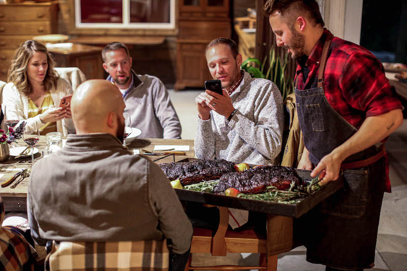 Group of five people gathered around a table, one person serving food, others using phones, in a cozy indoor setting.