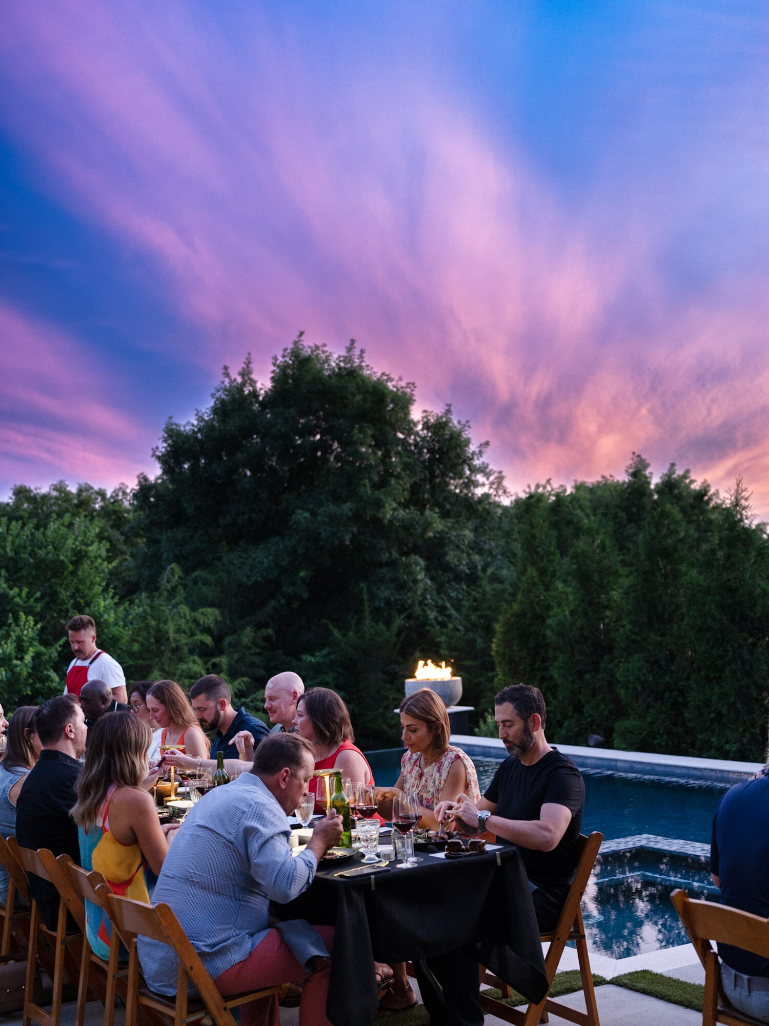 People dining outdoors at sunset with a colorful sky and lush trees in the background.