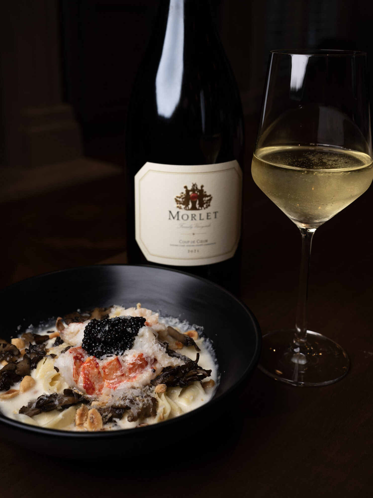 A bottle of Morlet wine, a glass of white wine, and a bowl of pasta with black truffle on a dark table.