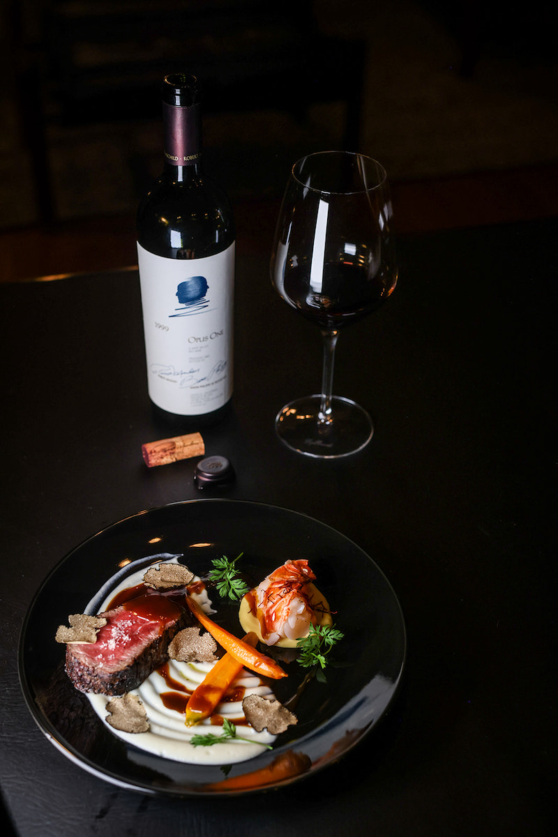 A black plate with cooked meat, vegetables, and garnishes, a bottle of wine, a glass of red wine, and a cork on a dark table.