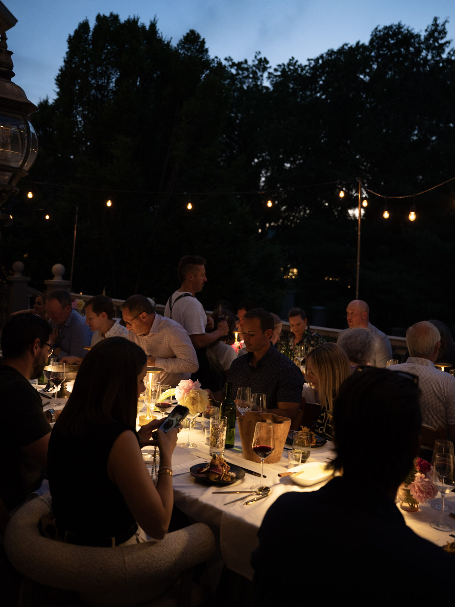 People dining outdoors at night with string lights and trees in the background, some using phones, warm lighting on table