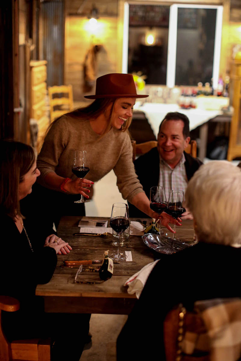 Group of people gathered around a table, woman standing and smiling, holding a glass of wine, in a cozy indoor setting.