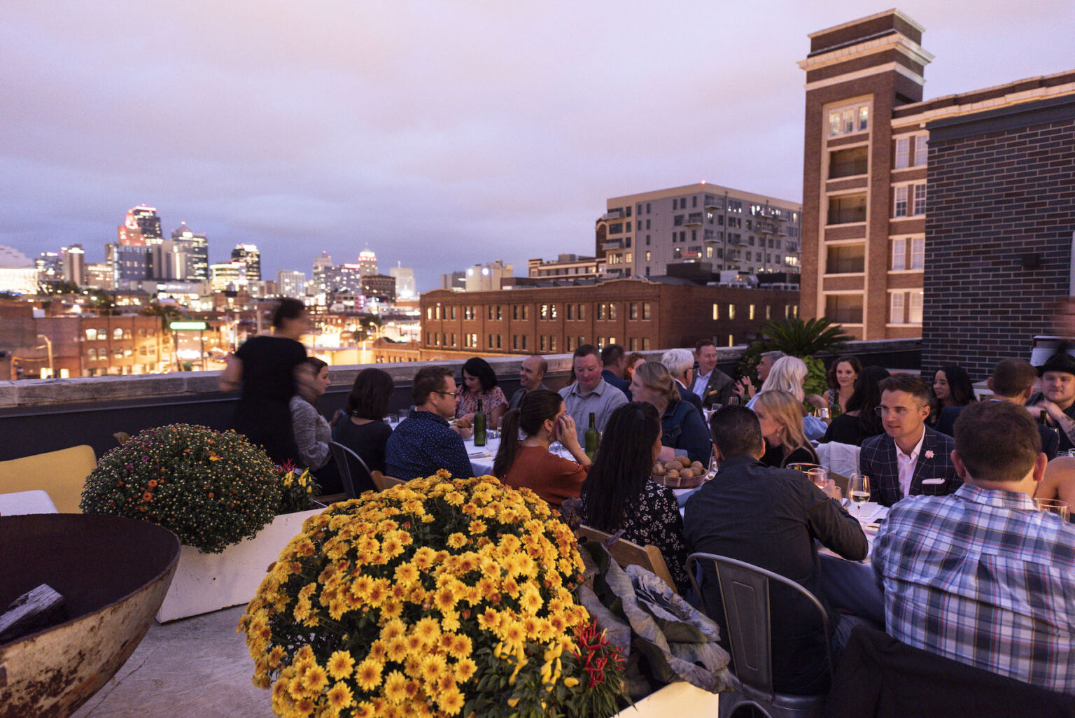 People dining on a rooftop terrace with city skyline in the background during evening, with yellow flowers in foreground.