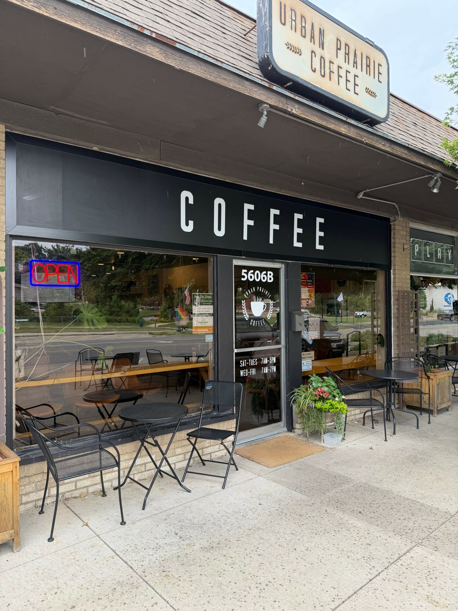 Coffee shop entrance with outdoor seating, sign, and menu display, viewed from the sidewalk.