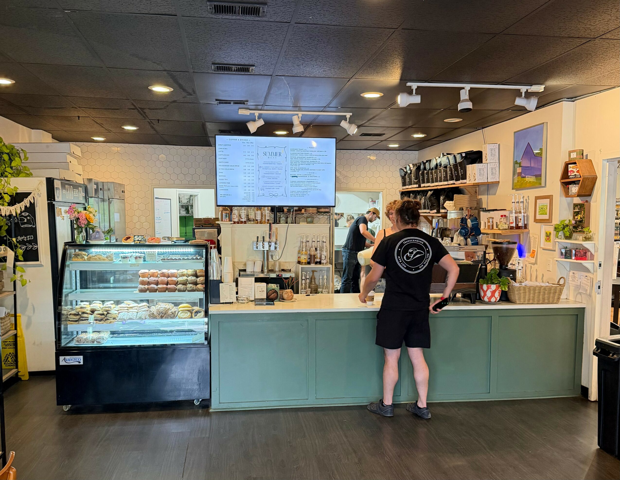 Person standing at a counter in a cafe, with a display case of desserts and a menu screen above. Bright interior.