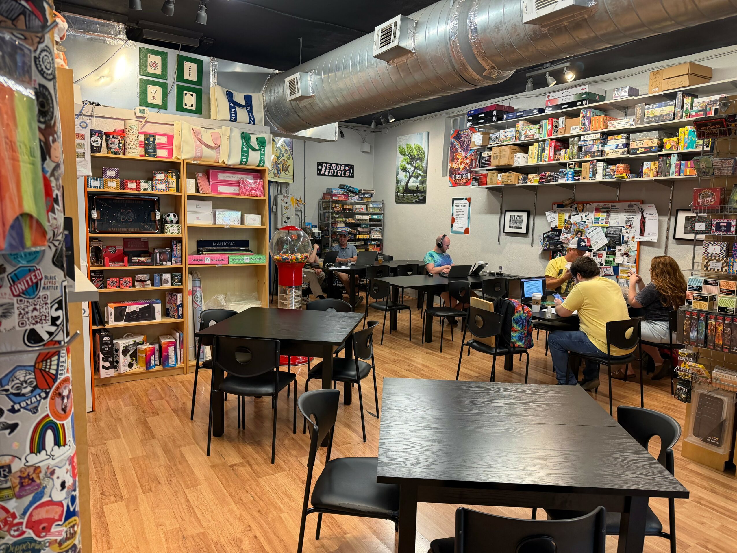 Indoor space with tables, chairs, bookshelves, and people reading or working, with a ceiling duct and wall decorations.