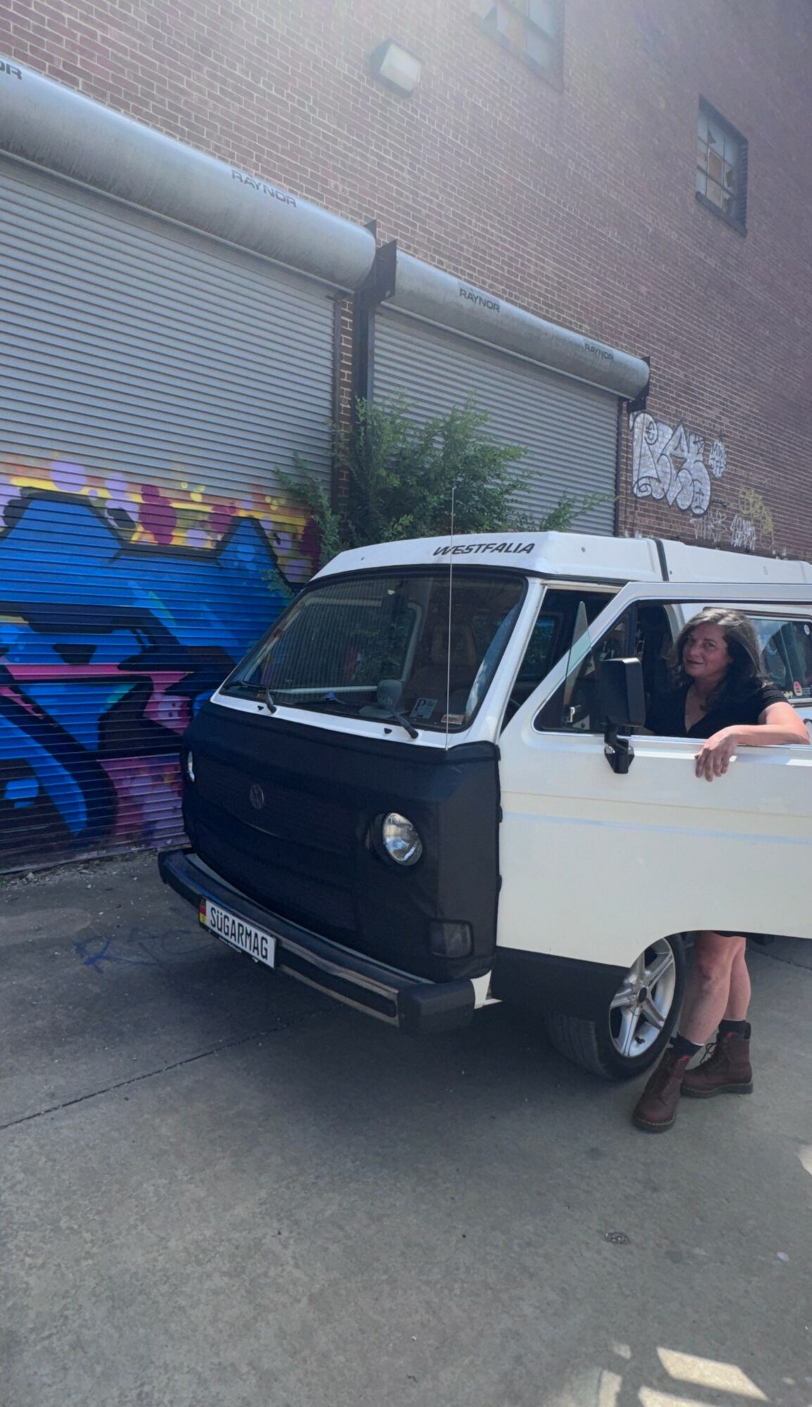 Person standing next to a white van with a woman inside, in front of a graffiti-covered wall.