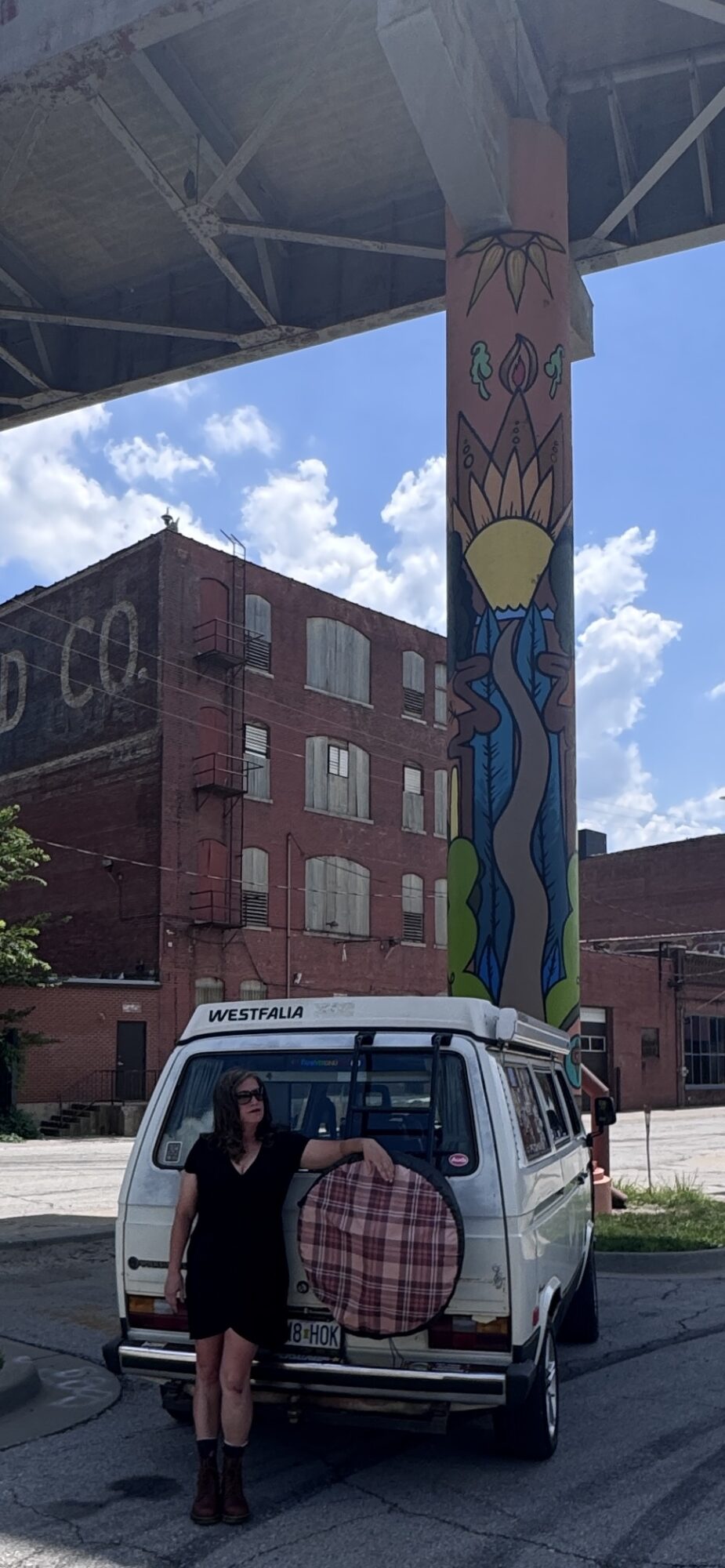 Person standing next to a vehicle with a heart-shaped design, under a colorful painted pillar and a bridge, brick building in background.