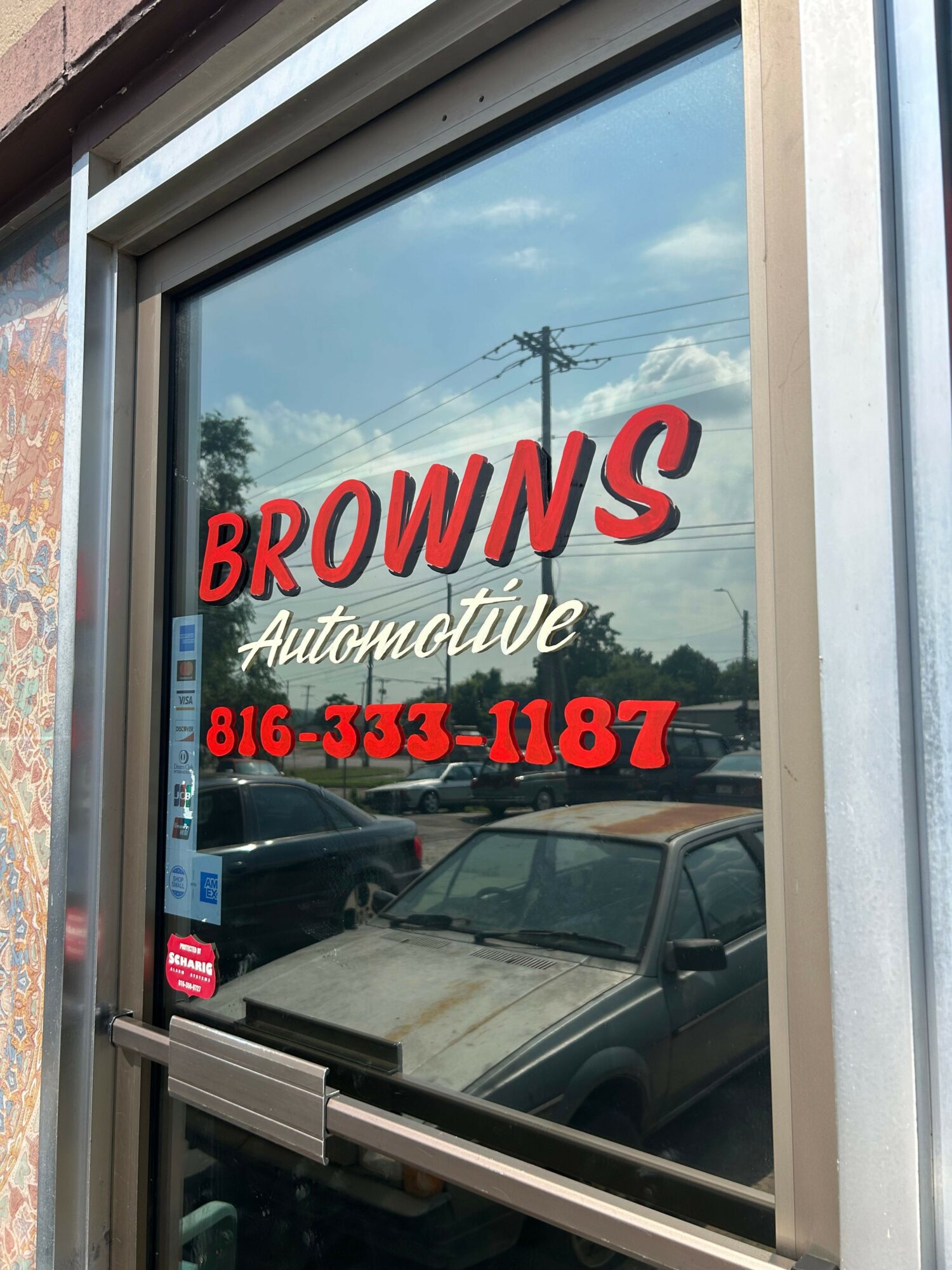 Store window with red and black text advertising BROWN'S Auto Body, phone number 816-636-6481, and a reflection of the sky and power lines.