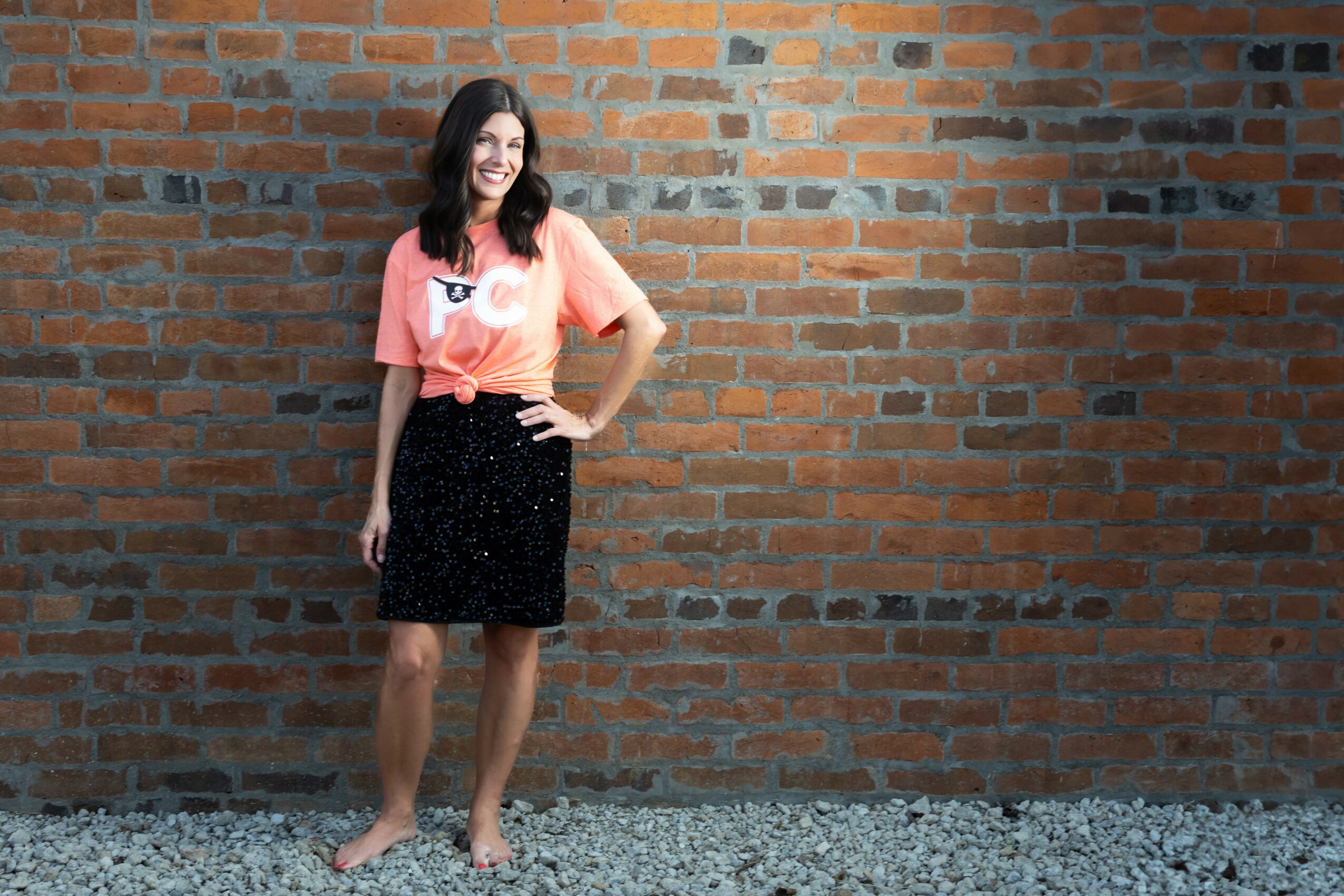 Young woman with long dark hair smiling, standing barefoot against a brick wall, wearing a pink T-shirt and black skirt.
