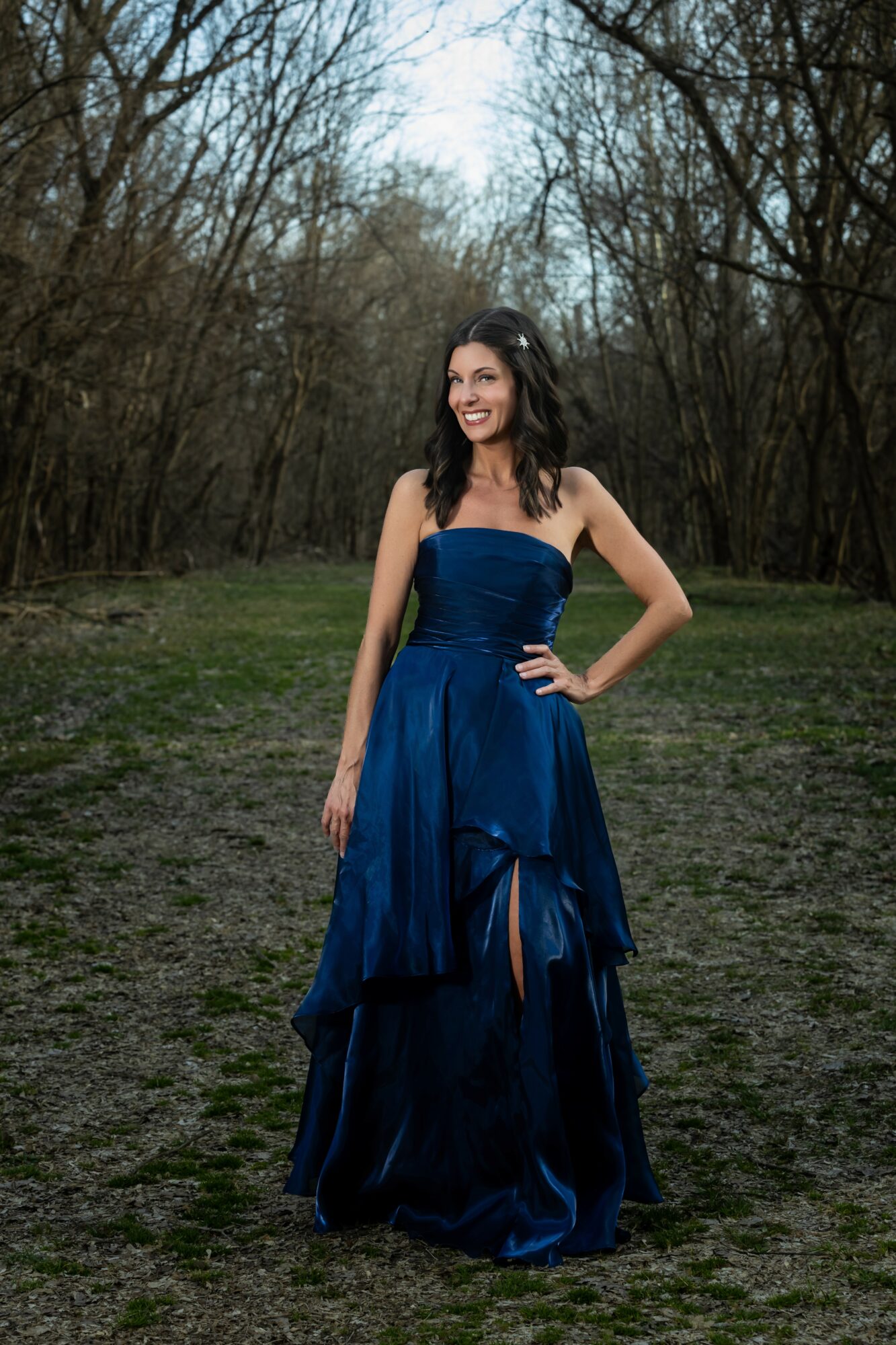 Young woman in a strapless blue gown standing outdoors in a wooded area, smiling, with hands on hips.