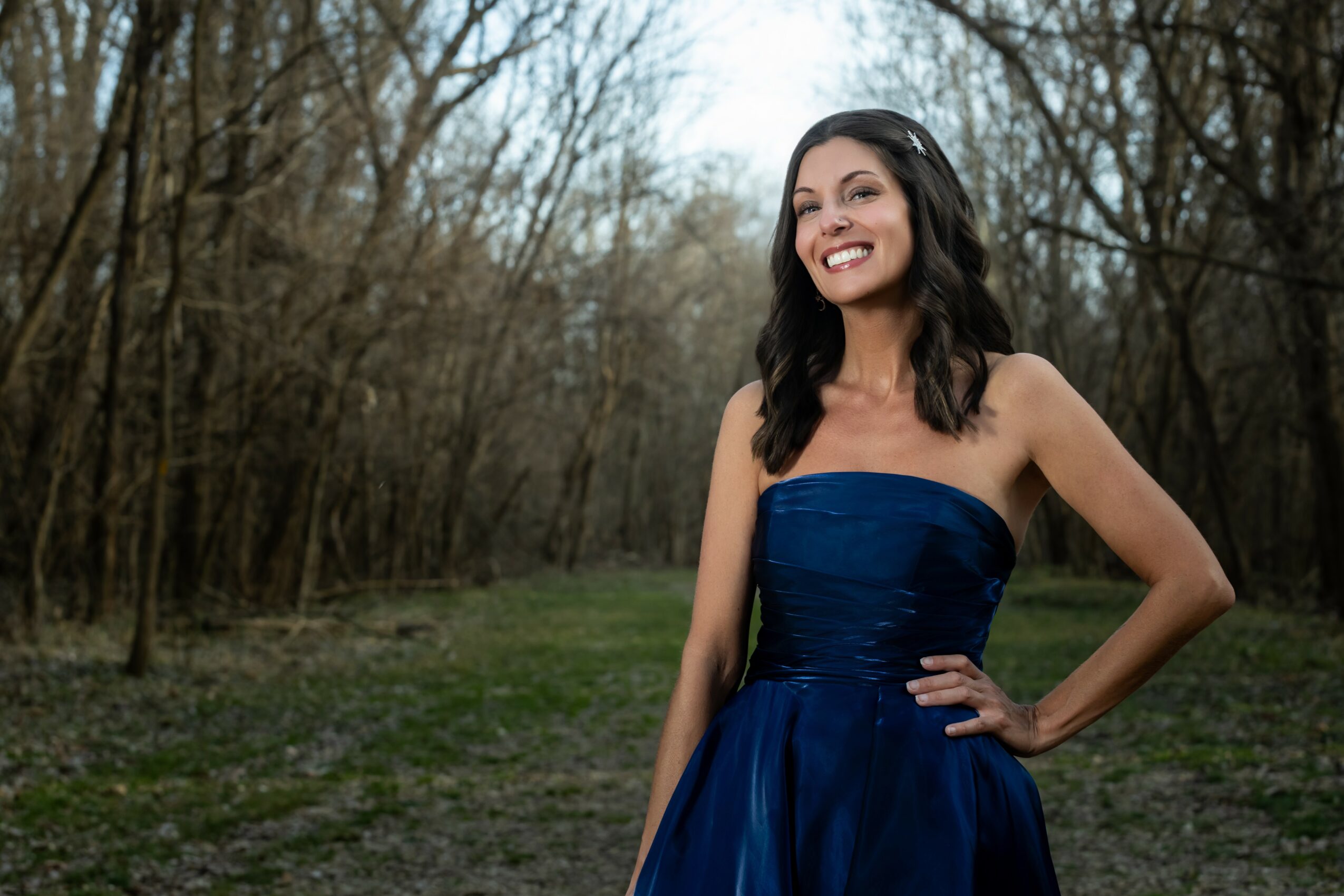 Smiling woman in a strapless blue dress outdoors with trees in the background.