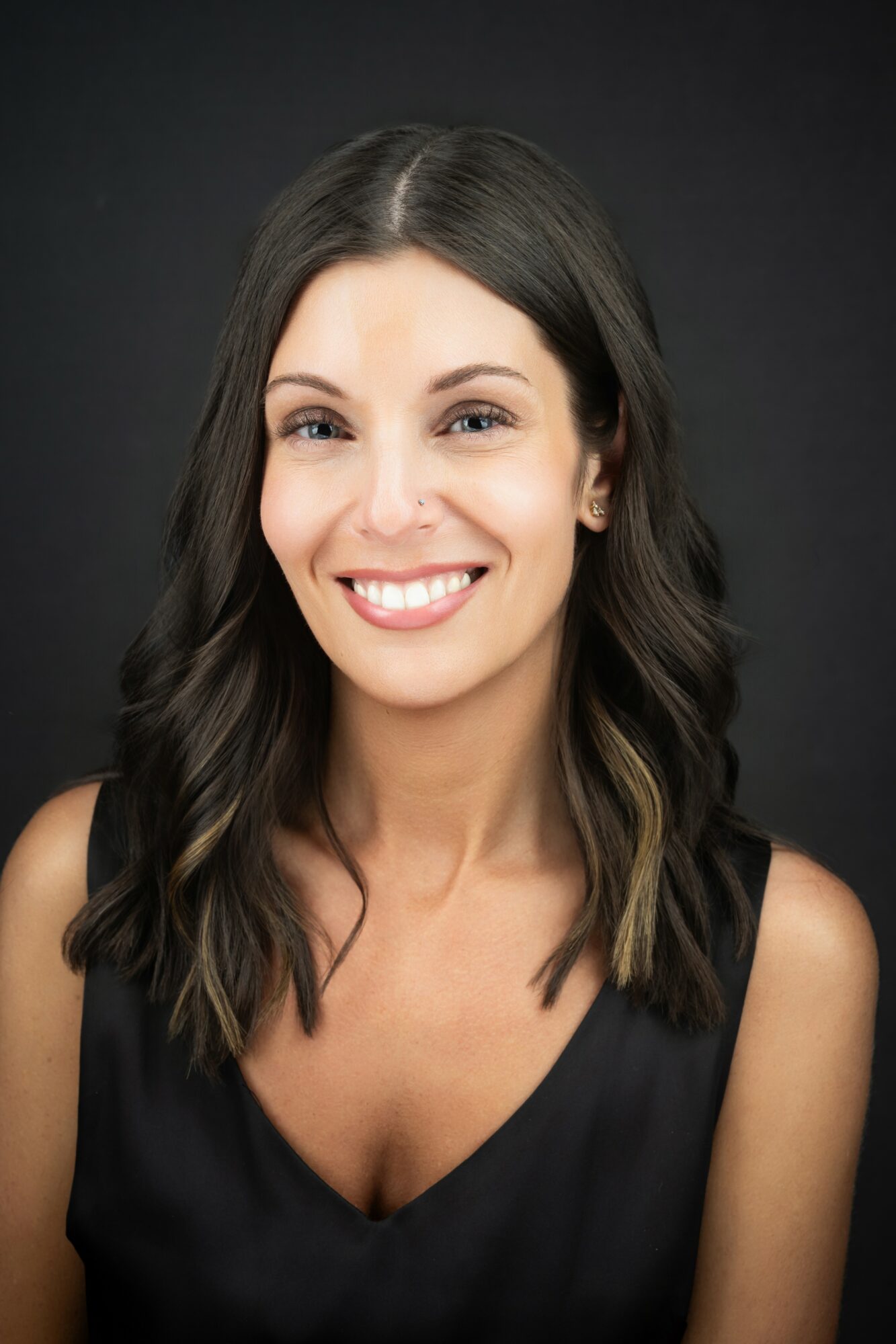 Young woman with long dark hair smiling, wearing a black sleeveless top, against a dark background.
