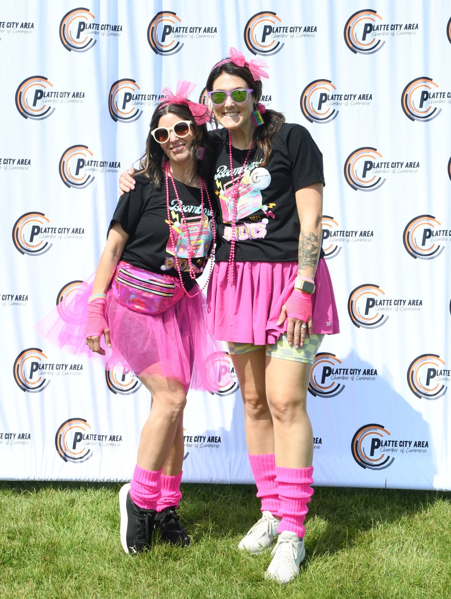 Two women in colorful costumes stand on grass in front of a backdrop with logos, smiling and posing for the photo.