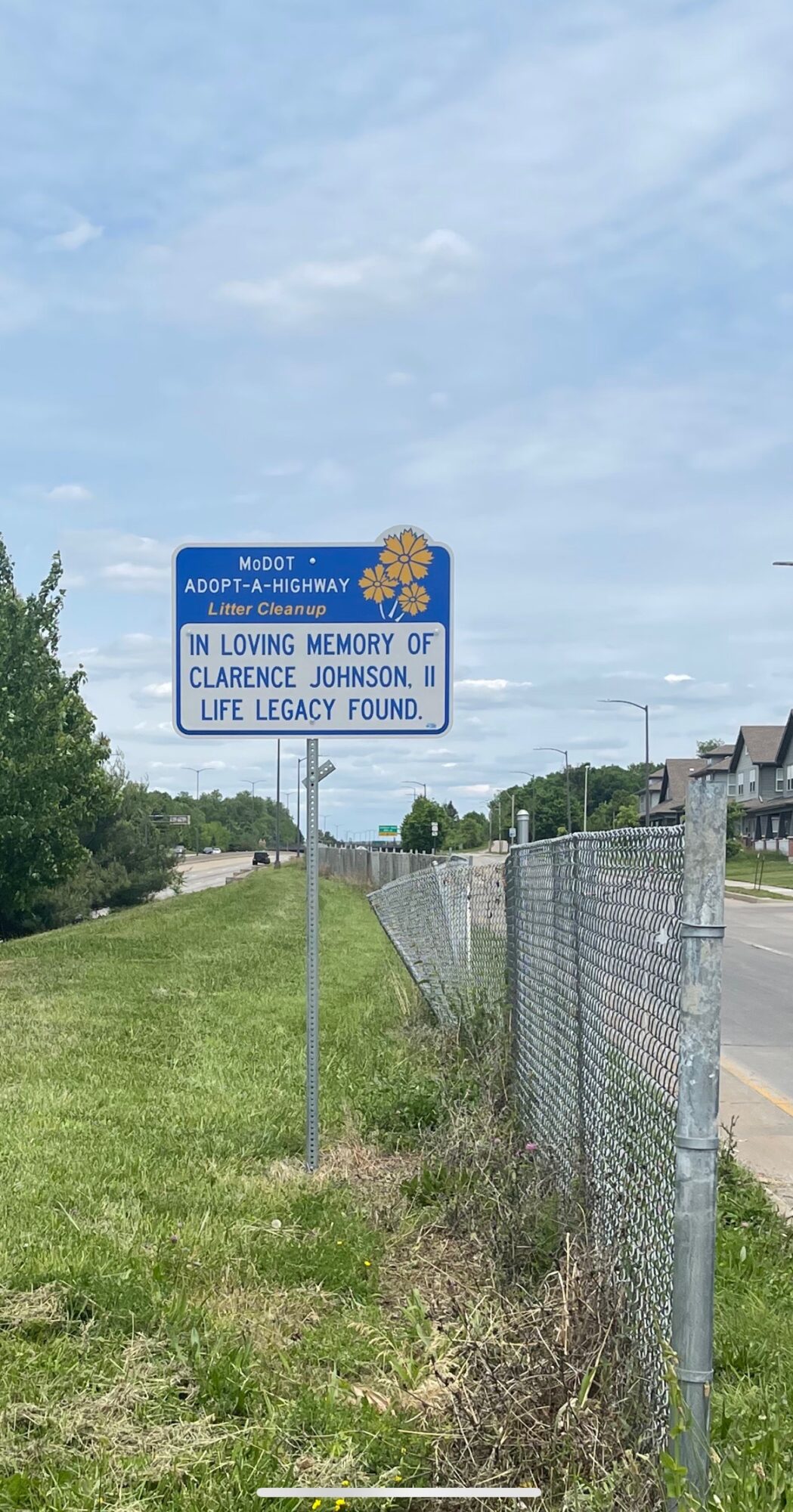 Blue memorial sign on grass beside a chain-link fence, with a cloudy sky above and a road to the right.