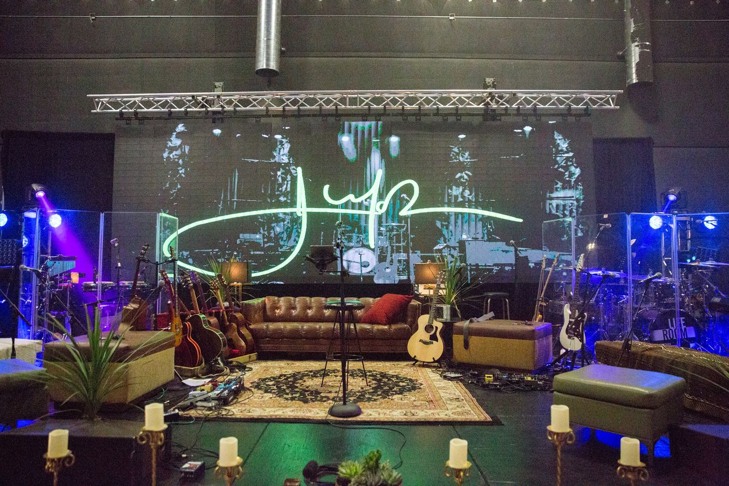Stage with a sofa, guitars, and candles, set for a performance with lighting and a large screen backdrop.