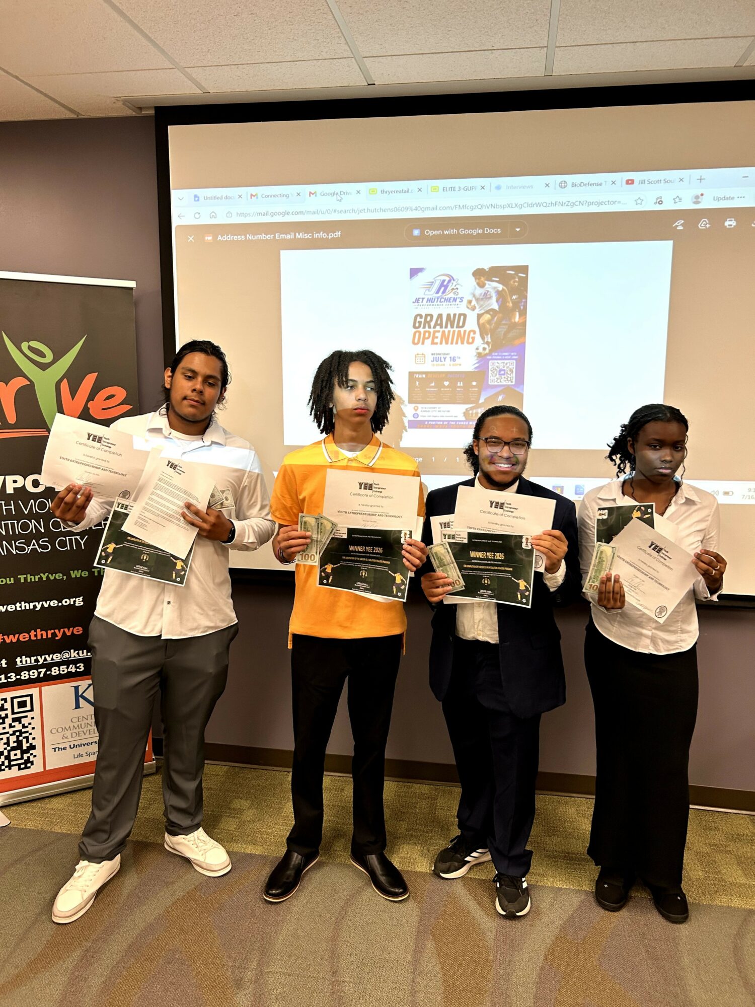 Four people stand in front of a presentation screen holding certificates and newspapers, in a room with a banner on the left.