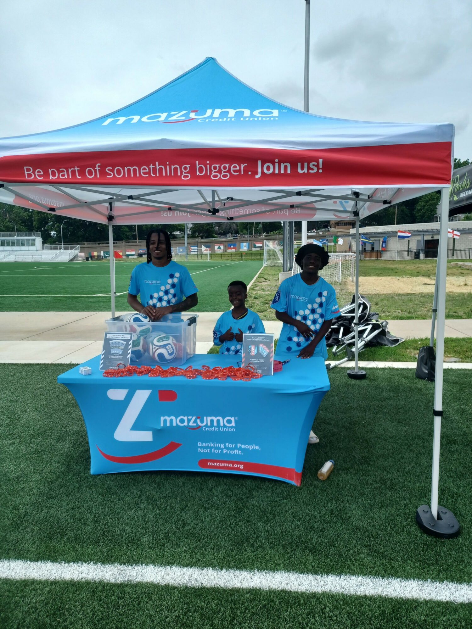 Two people in blue shirts stand behind a promotional table with a blue canopy and a Z Mountain banner, outdoors on grass.