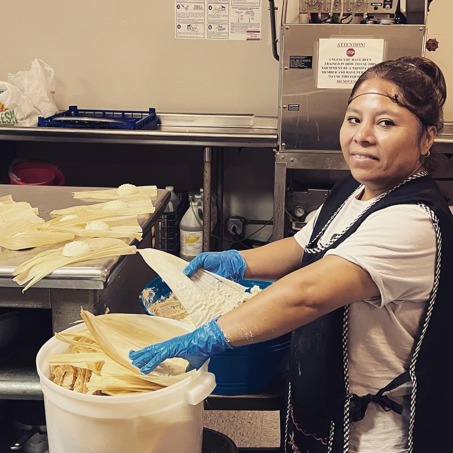 Woman wearing gloves and apron handling food in a kitchen, with stacked food items on a table behind her.